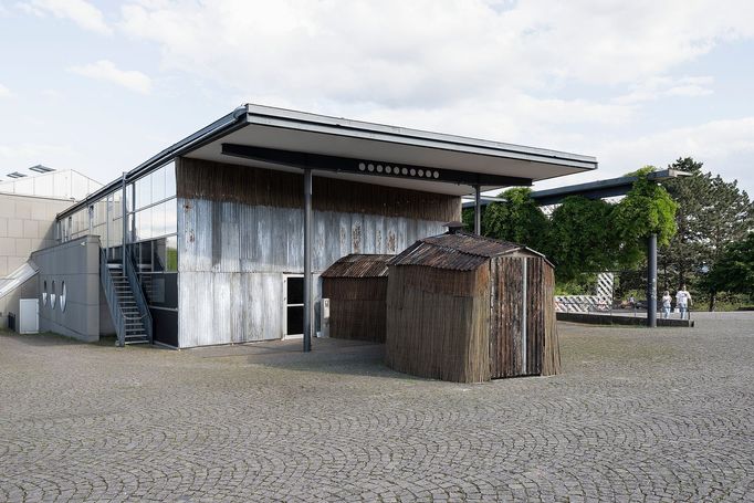 Do výstavního pavilonu Documenta Halle lidé vstupují temným tunelem, kde svítí petrolejové lampy a zdmi rachotí zvuk projíždějícího vlaku.