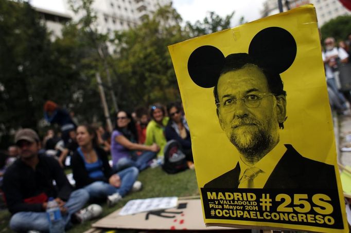 Demonstrators sit next to a sign with an image of Spanish Prime Minister Mariano Rajoy as they wait for the start of an assembly before an anti-austerity demonstration in Madrid September 25, 2012. Police prepared on Tuesday for anti-austerity demonstrations in Spain's capital ahead of the government's tough 2013 budget that will cut into social services as the country teeters on the brink of a bailout. The sign reads, "Occupy Congress, 25S, Madrid and Valladolid". REUTERS/Susana Vera (SPAIN - Tags: BUSINESS CIVIL UNREST) Published: Zář. 25, 2012, 2:25 odp.