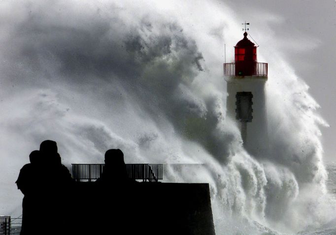 Two silhouetted people watch a powerfull wave as it hits a lighthouse at the entrance of Les Sables D'Olonne harbour.