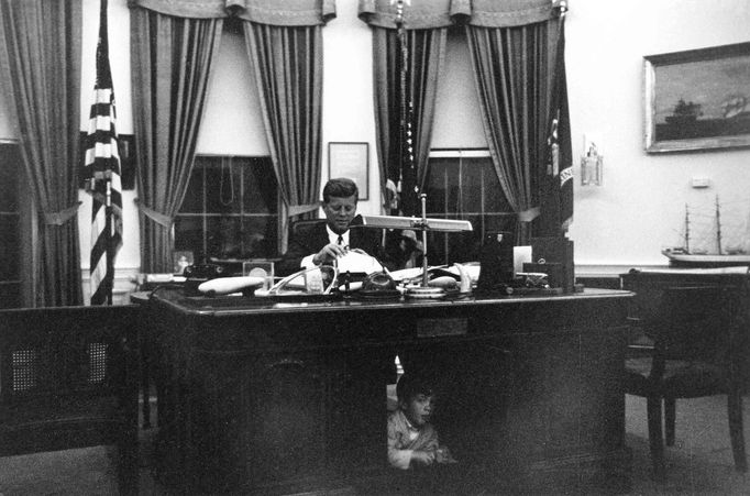 Former United States President John F. Kennedy sits as his desk in the Oval Office while his son, John F. Kennedy Jr., looks out from underneath, at the White House in Washington in this handout image taken on October 17, 1963.