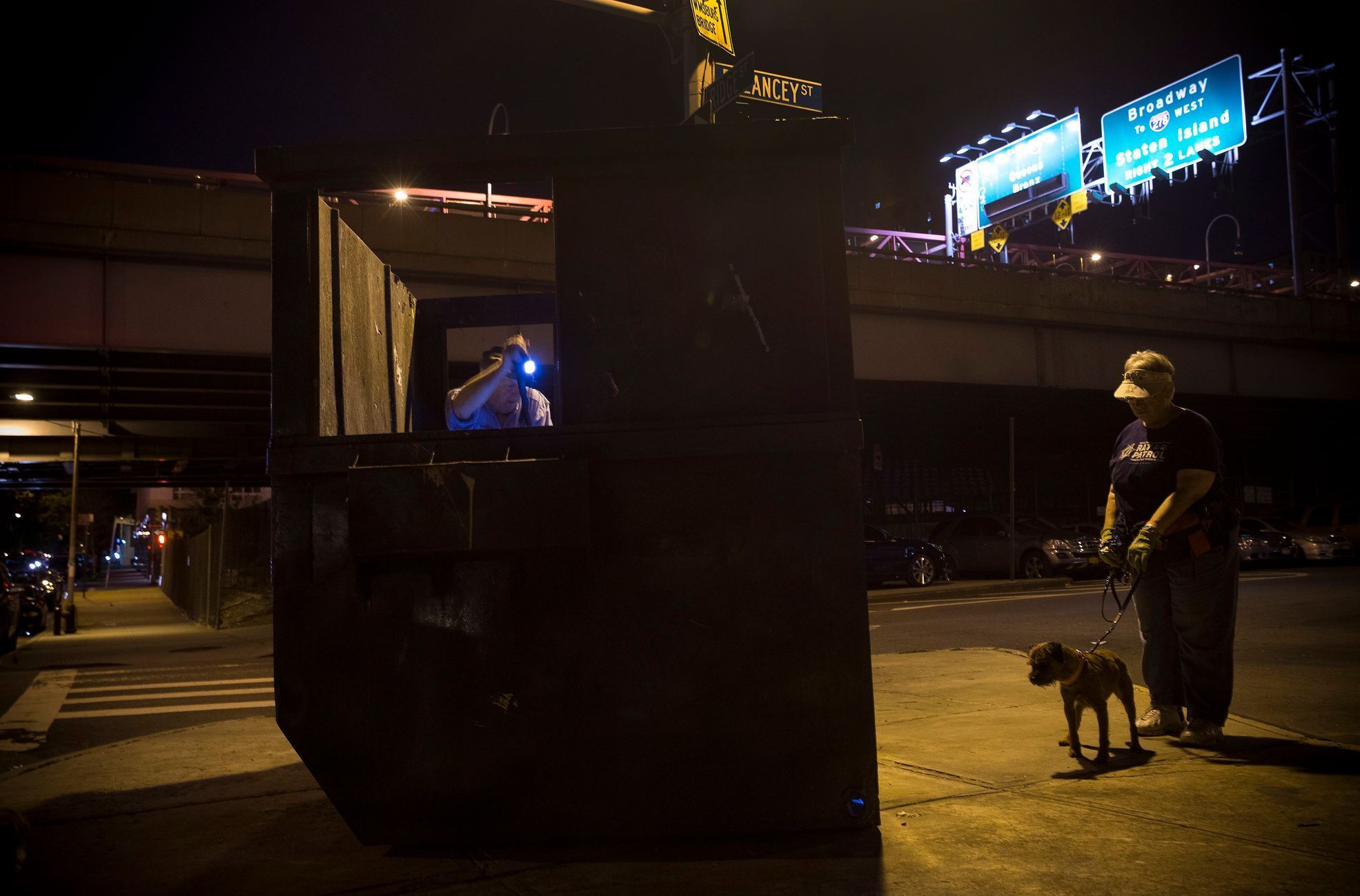 Reynolds of the Ryders Alley Trencher-fed Society, uses a flashlight to look for rats in a dumpster as Judy and her Border Terrier Merlin stand by, ready to make a kill, during organized rat hunt on N